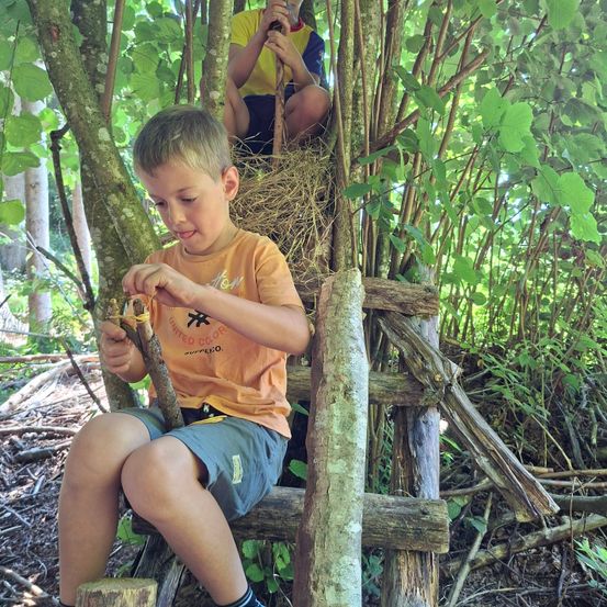 Ein kleiner Junge sitzt auf einer Bank aus Stöcken im Wald, hält einen Stock in der Hand, während ein anderes Kind in einem Baumhaus sitzt.