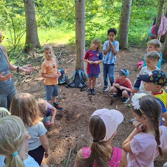 Eine Gruppe von Kindern ist in einer Waldlichtung versammelt. Eine Frau spricht zu ihnen, während einige Kinder auf dem Boden sitzen. Ein Junge steht und andere schauen aufmerksam zu ihr.
