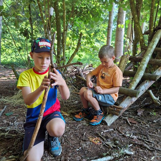 Zwei Jungen im Wald, einer hält einen Stock, der andere sitzt auf einer Holzleiter. Beide tragen sommerliche Freizeitkleidung.