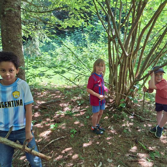 Drei Kinder stehen in einem Wald und halten Stöcke. Eines trägt ein Argentinien-Trikot, ein anderes ein rotes und das dritte ein rosa Trikot.