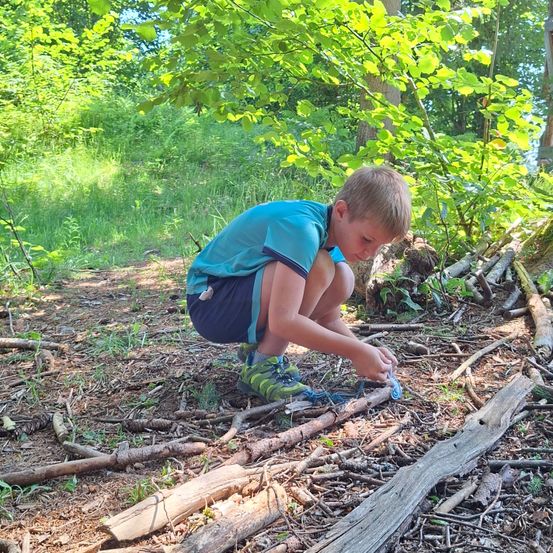 Ein junger Junge kniet auf einem Waldpfad und knotet eine blaue Schnur. Der Pfad ist von Bäumen und Grün umgeben.