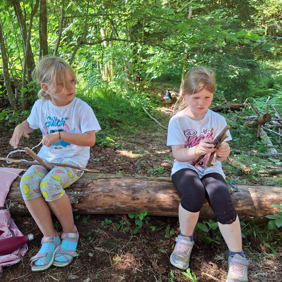 Zwei junge Mädchen sitzen auf einem Baumstamm im Wald. Eines hält einen Stock, das andere ein Stück Holz. Sie sind von Grün umgeben und es gibt einen Rucksack.