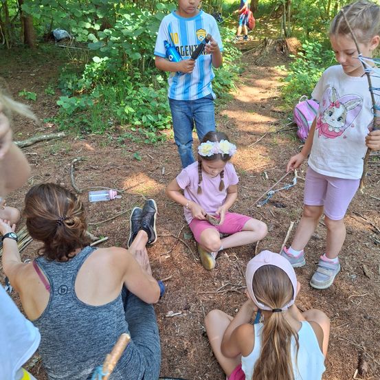 Mehrere Kinder in einer Waldumgebung, eine mit einem Stock, eine andere in rosa Shorts mit einem Gegenstand, und eine Frau in Grau sitzt auf dem Boden.