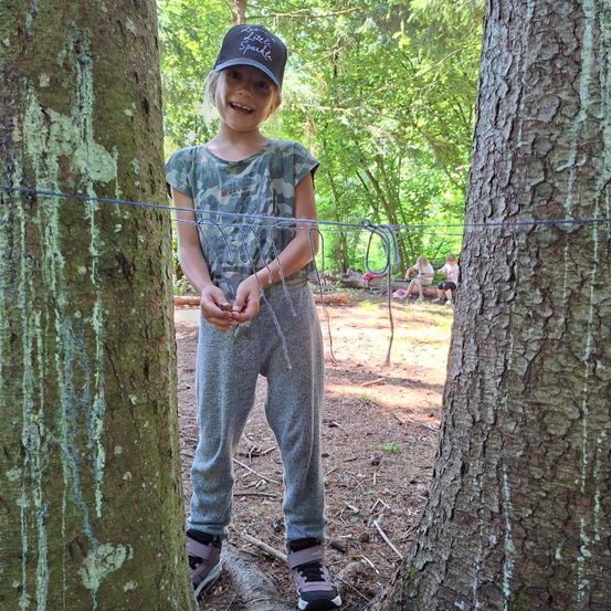 Ein kleines Mädchen in Camouflage-Kleidung und einer Baseball-Kappe steht zwischen zwei Bäumen und hält ein Kabel. Sie scheint zu lächeln und posiert für ein Foto. Hinter ihr sitzen und stehen eine Gruppe von Menschen auf dem Boden.