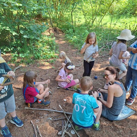 Eine Gruppe von Kindern und Erwachsenen befindet sich in einem Wald, einige stehen und andere sitzen auf dem Boden. Sie halten Stöcke und scheinen eine Aktivität zu machen. Eine Frau mit Sonnenbrille sitzt auf einer Matte und beobachtet die Kinder.