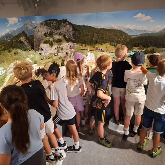 Eine Gruppe von Kindern und Teenagern steht vor einer großen Wandmalerei und beobachtet eine Modelleisenbahn-Aufstellung an der Wand. Die Wandmalerei zeigt eine bergige Landschaft mit Tunneln und einer Kirche.