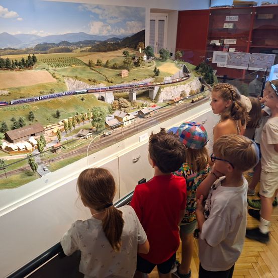 Eine Gruppe von Kindern beobachtet ein Miniatur-Zugmodell mit einer Modellstadt und Bergen im Hintergrund. Die Kinder stehen vor einer Glasvitrine.
