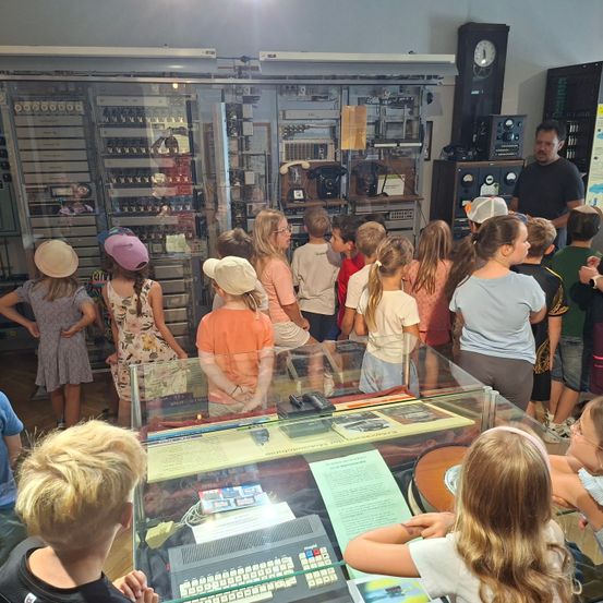 Eine Gruppe von Kindern und Erwachsenen versammelt sich um eine Glasvitrine mit alter Technologie. Sie tragen Hüte und einige haben Brillen. Ein Mann erklärt ihnen etwas.