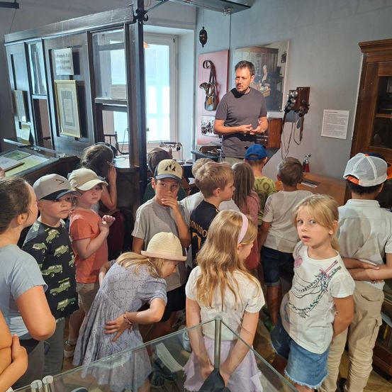 Ein Mann erklärt etwas einer Gruppe von Kindern in einem Raum mit verschiedenen Objekten und einer Glasvitrine.