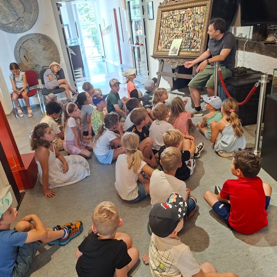 Ein Mann steht vor einer Gruppe von Kindern in einem Raum. Sie sitzen auf dem Boden und hören ihm zu. Auf dem Tisch vor ihm steht ein gerahmtes Kunstwerk.