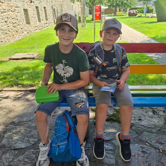 Zwei junge Jungen sitzen auf einer bunten Bank im Freien. Einer hält eine grüne Lunchbox und der andere liest ein Buch. Sie tragen Freizeitkleidung und Mützen. Hinter ihnen befindet sich eine Steinmauer und ein Rasenbereich mit einem Weg.