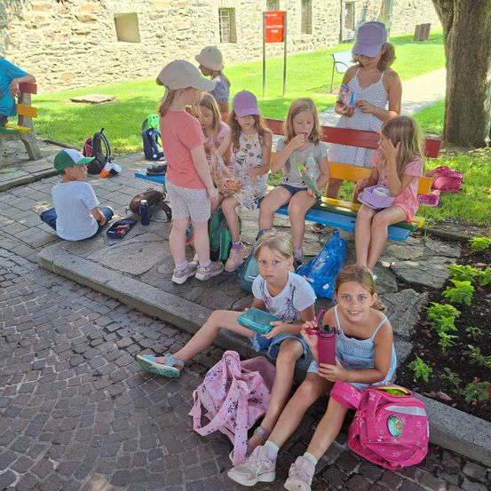 Eine Gruppe junger Kinder, einige mit Mützen, sitzen auf Bänken und einer Stufe, möglicherweise in einem Park. Einige essen, während andere Taschen und Flaschen um sich herum haben.