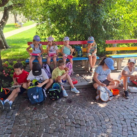 Mehrere Kinder sitzen auf einer Bank, einige haben Essen und Snacks. Einige der Kinder sitzen auf dem Boden.