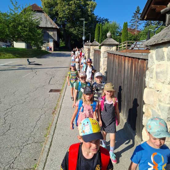Eine Reihe von Kindern mit Mützen und Rucksäcken geht eine Straße entlang. Im Hintergrund sind Bäume und ein Haus zu sehen.