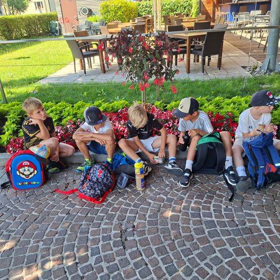 Fünf Jungen sitzen auf dem Boden, einige tragen Mützen, alle mit Rucksäcken. Sie scheinen zu warten. Hinter ihnen sind Tische und Stühle, ein Garten mit Blumen und ein Zaun.