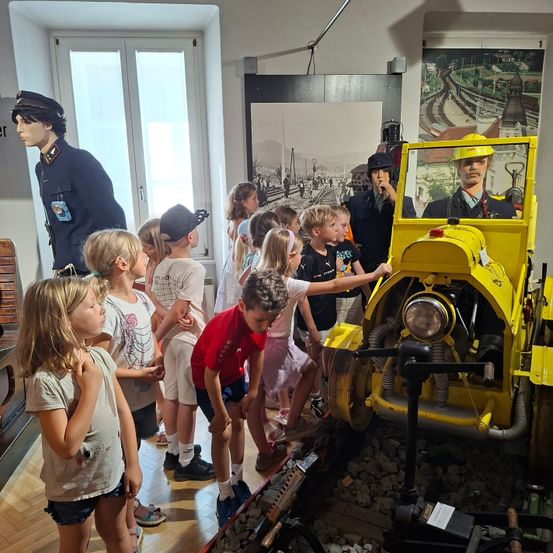 Kinder erkunden ein gelbes Fahrzeug in einem Museum, mit in Uniformen gekleideten Mannequins in der Nähe. Mannequins und historische Bilder sind im Hintergrund zu sehen.