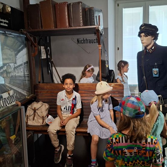 Kinder sitzen auf einer Bank in einem Museum, ein Kind trägt einen Hut. Ein Dummy in Uniform steht hinter ihnen, und Taschen und Koffer befinden sich auf einem Regal. Ein großer Glaskasten ist links.
