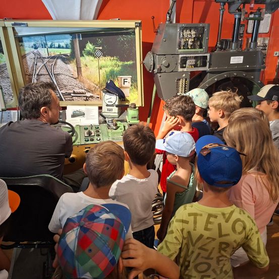 Eine Gruppe von Kindern beobachtet einen Mann, der einen Zugsimulator bedient, wobei eine Eisenbahnstrecke auf einem Monitor angezeigt wird. Der Raum hat rote Wände und mechanische Geräte.