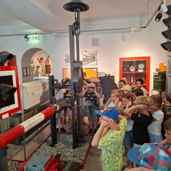 Eine Gruppe von Kindern erkundet eine Eisenbahnausstellung, steht um eine alte Signalbox und Maschinen herum, wobei ein Kind sich gegen einen rot-weißen Pfosten lehnt.