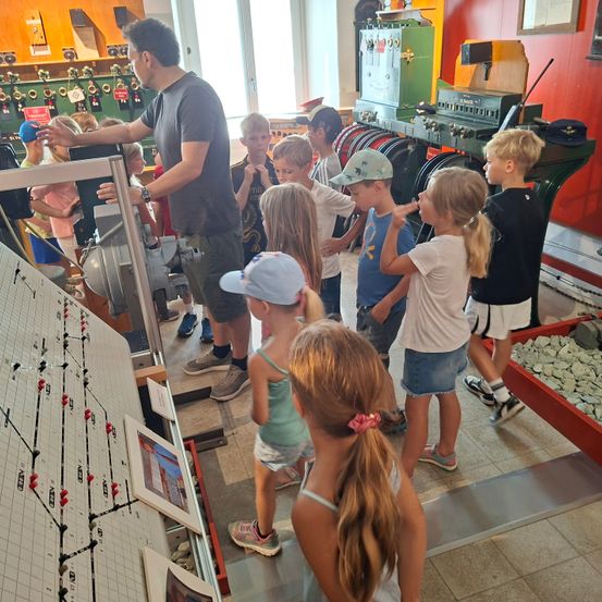 Ein Mann demonstriert eine Maschine einer Gruppe von Kindern in einem Raum mit verschiedenen Maschinen und einem Fenster, das das Freie zeigt.