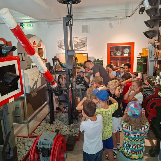 Eine Gruppe von Kindern und ein erwachsener Mann betrachten eine Ausstellung in einem Museum, umgeben von Maschinen und Zahnrädern.