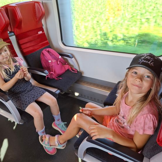 Zwei junge Mädchen in einem Zug, eine mit einem rosa Rucksack und die andere mit einer schwarzen Kappe. Sie sitzen mit einem Fenster dahinter, das grüne Bäume zeigt.