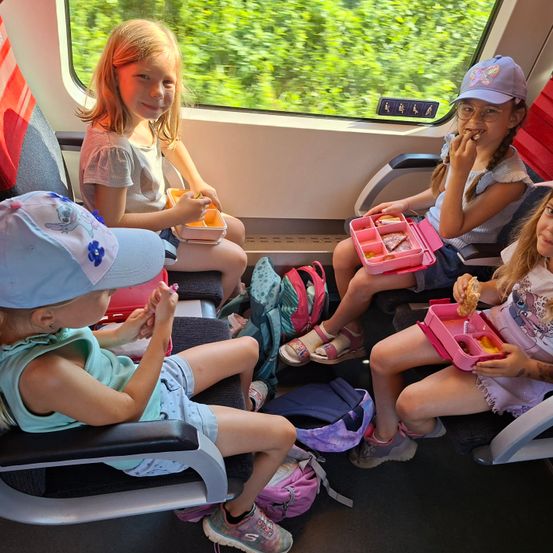 Vier junge Mädchen sitzen in einem Zug und essen aus ihren Brotdosen. Zwei tragen Mützen und die anderen haben eine Brille. Im Fenster draußen sind Bäume zu sehen.
