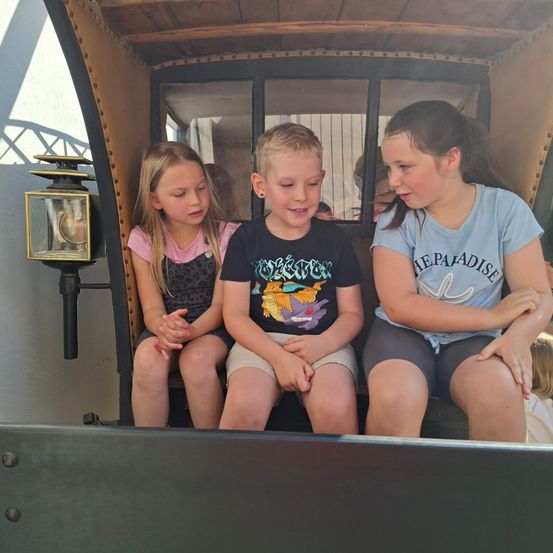 Drei Kinder sitzen in einer alten Kutsche, mit zwei Mädchen und einem Jungen, alle schauen neugierig nach vorne. Das Mädchen rechts trägt ein blaues T-Shirt mit weißen Buchstaben.
