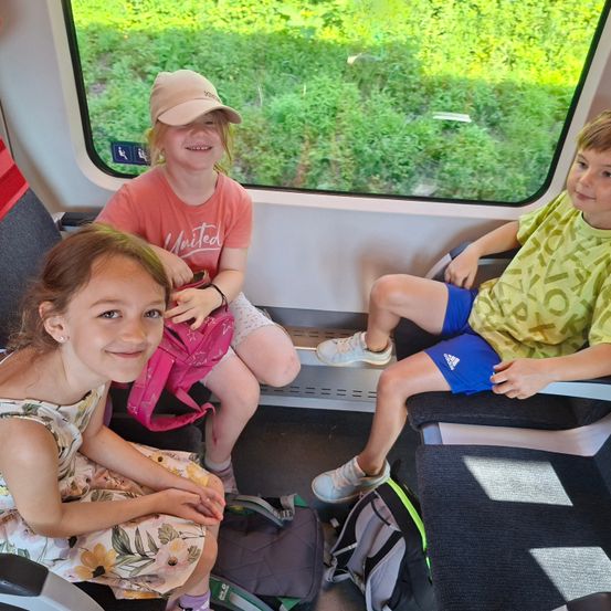Drei Kinder sitzen in einem Zug. Ein Mädchen lächelt zur Kamera, neben ihr steht ein Rucksack. Ein weiteres Mädchen sitzt hinter ihr mit einem rosa Rucksack. Ein Junge sitzt neben ihr.