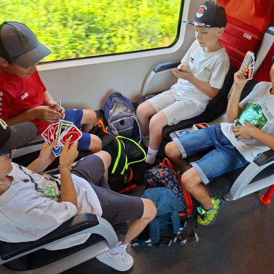 Vier Jungen sitzen in einem Zug und spielen Karten. Einer hält ein Kartenspiel, ein anderer benutzt sein Handy. Rucksäcke und ein rotes Gepäcketikett sind sichtbar.