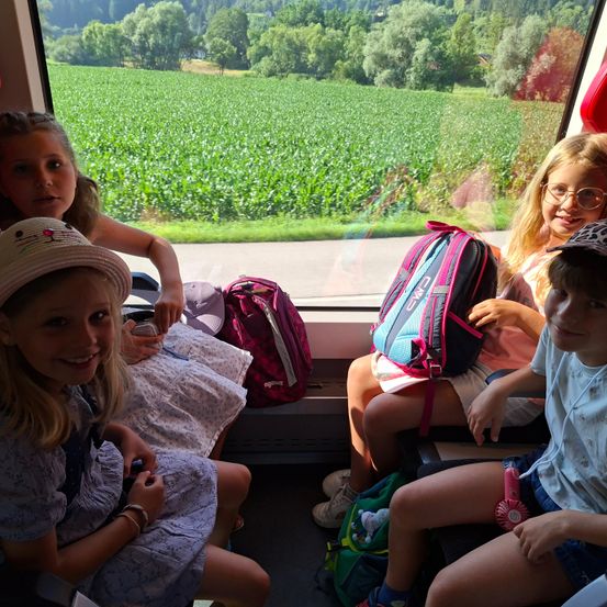 Vier Kinder sitzen in einem Zug, lächeln und schauen aus dem Fenster. Ein Mädchen trägt einen Katzenhut. Das Feld und die Bäume draußen sind durch das Fenster zu sehen.