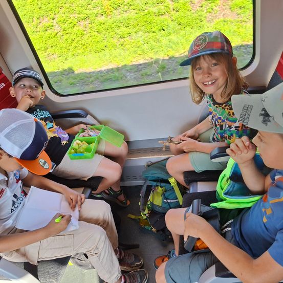 Vier Kinder sitzen in einem Zug, jeder mit einem Essenstablett. Sie alle sehen glücklich aus. Durch das Fenster ist grünes Gelände zu sehen.