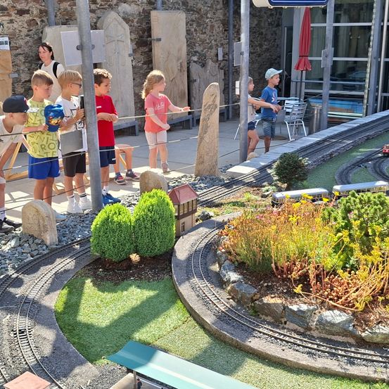 Eine Gruppe von Kindern und Erwachsenen steht in einem Gartenbereich mit Miniatur-Bahnschienen, Pflanzen und einem kleinen Haus. Einige halten an den Zügen befestigte Schnüre.