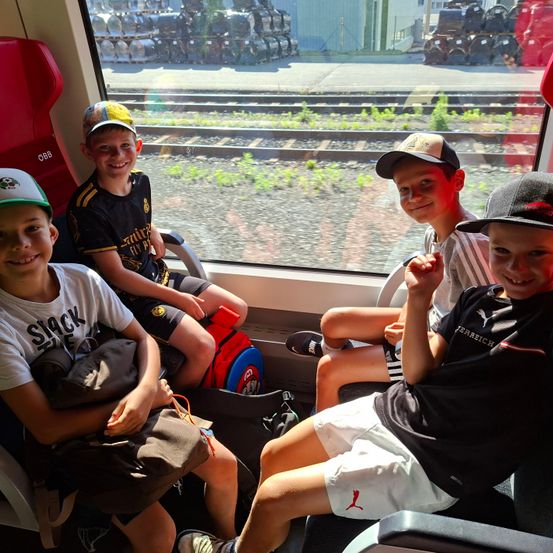 Vier Jungen sitzen in einem Zug, dem Kameramann zugewandt. Jeder trägt einen Baseball-Cap. Einer hält eine schwarze Tasche, ein anderer einen roten und blauen Rucksack. Das Fenster zeigt Gleise und die Ansicht einer Fabrik.