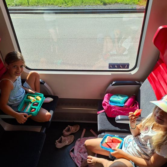 Zwei junge Mädchen sitzen in einem Zug, eine isst Wassermelone und die andere hält eine Lunchbox mit verschiedenen Snacks. Der Zug hat ein Fenster, das eine Straße draußen zeigt.