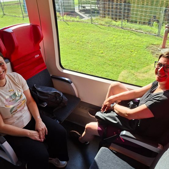 Zwei Frauen sitzen in einem Zug und lächeln. Eine trägt eine Brille, die andere ein weißes T-Shirt. Ein roter Sitz befindet sich hinter der Frau im weißen T-Shirt. Der Blick nach draußen zeigt grünes Gras und Gebäude.