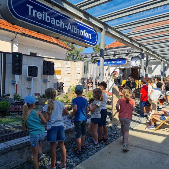 Kinder stehen auf einer Schotterplattform vor einem Modelleisenbahnbahnhof. Der Bahnhof hat ein blaues Schild mit der Aufschrift Treibach-Althofen. Einige Kinder sitzen auf einer Bank.