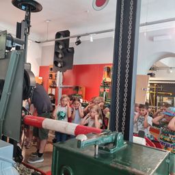 Eine Gruppe von Kindern mit Erwachsenen steht vor einer Ampel und einer Zugausstellung. Die Kinder decken ihre Ohren ab, als wären sie überrascht. Die Umgebung hat rote Wände und industrielle Elemente.