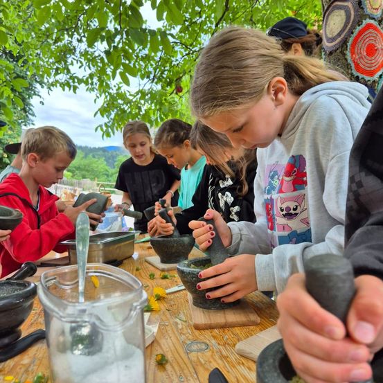 Kinder versammeln sich um einen Tisch im Freien und mahlen mit Mörsern und Stößeln etwas. Einige haben Blumen auf ihren Tischen.