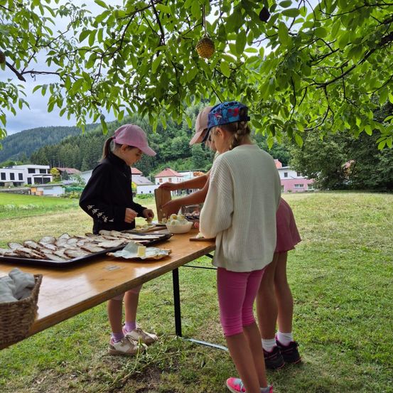 Drei Mädchen stehen bei einem Picknicktisch mit Essen unter einem Baum. Ein Mädchen trägt einen rosa Hut und rosa Shorts.
