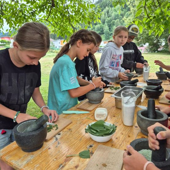 Mehrere Kinder sind um einen Holztisch versammelt, wobei jedes mit einem Mörser und Stößel Blätter in eine Schüssel zermahlt. Der Tisch ist mit verschiedenen Zutaten gefüllt, und sie befinden sich in einem Grasbereich mit Bäumen.