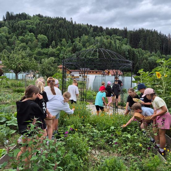 Eine Gruppe von Kindern arbeitet in einem Garten, einige pflücken Blumen, andere kümmern sich um Pflanzen. Ein Pavillon und ein Gebäude mit Gewächshaus sind im Hintergrund zu sehen.