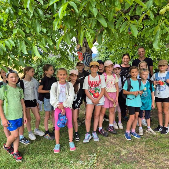 Eine Gruppe von Kindern und Erwachsenen steht unter einem großen Baum, trägt Sommerkleidung und Sneakers, wobei ein Kind einen Rucksack und ein anderes Kind eine Blume hält.