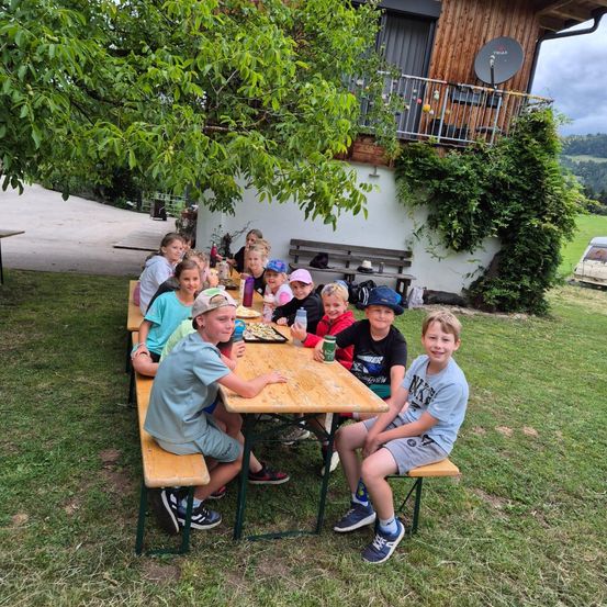 Eine Gruppe von Kindern isst draußen an einem Picknicktisch. Sie haben Snacks und Getränke auf dem Tisch. Ein Haus ist hinter ihnen.