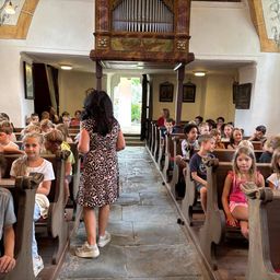 Eine Frau steht vor einem Kirchengang, mit einer Gruppe von Kindern auf beiden Seiten, die auf Kirchenbänken sitzen. Die Kirche hat Holzbänke, einen hölzernen Altar und eine kunstvolle Orgel.
