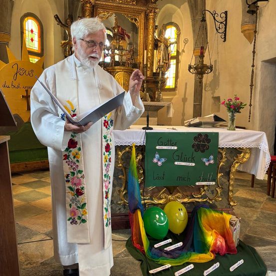 Ein älterer Mann, möglicherweise ein Priester, steht an einem Tisch in einer Kirche. Er hält ein Buch und gestikuliert mit der Hand. Der Tisch ist mit Luftballons, einem grünen Tuch und einem Schild mit der Aufschrift 'Gottes Glück' dekoriert. Hinter ihm befindet sich ein Altar mit Blumen und einer Statue von Jesus.