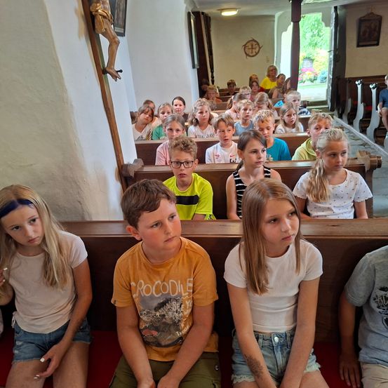Eine Gruppe von Kindern sitzt auf hölzernen Kirchenbänken, wobei einige fokussiert erscheinen und andere wegschauen. Eine Statue eines Heiligen ist an der Wand hinter ihnen angebracht.