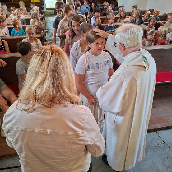 Ein Priester segnet eine Gruppe von Kindern in einer Kirche, eine Frau beobachtet von der Seite. Die Kinder sind lässig gekleidet, und der Priester trägt eine weiße Robe.
