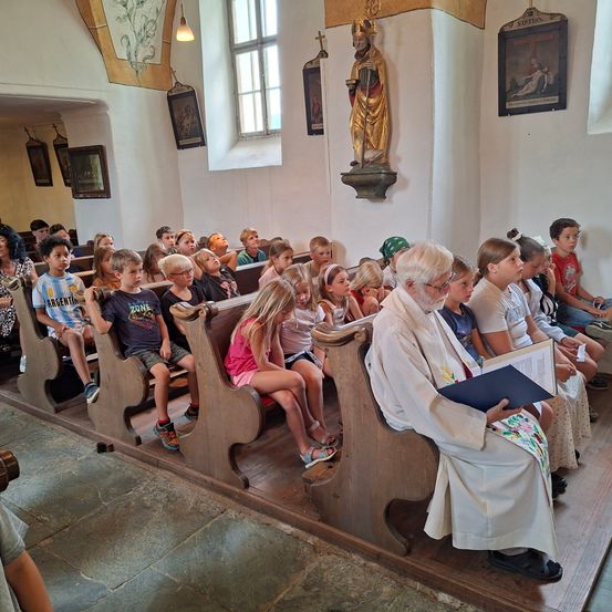 Ein Priester liest aus einem Buch in einer Kirche, umgeben von Kindern auf hölzernen Kirchenbänken. Eine goldene Statue und religiöse Gemälde schmücken die Wände.