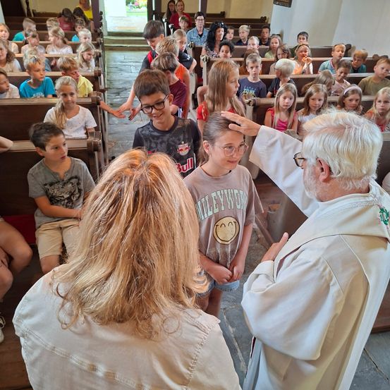 Ein Priester segnet ein Mädchen in einer Kirche, umgeben von Kindern, die auf Holzbänken sitzen. Der Priester trägt eine Brille und eine weiße Robe.